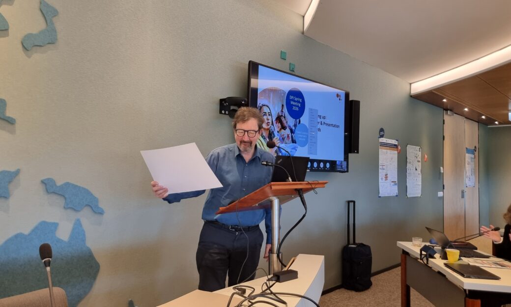 Prof. Creton stands with microphone in front of screen, holding a paper
