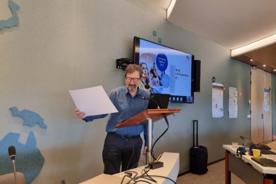 Prof. Creton stands with microphone in front of screen, holding a paper